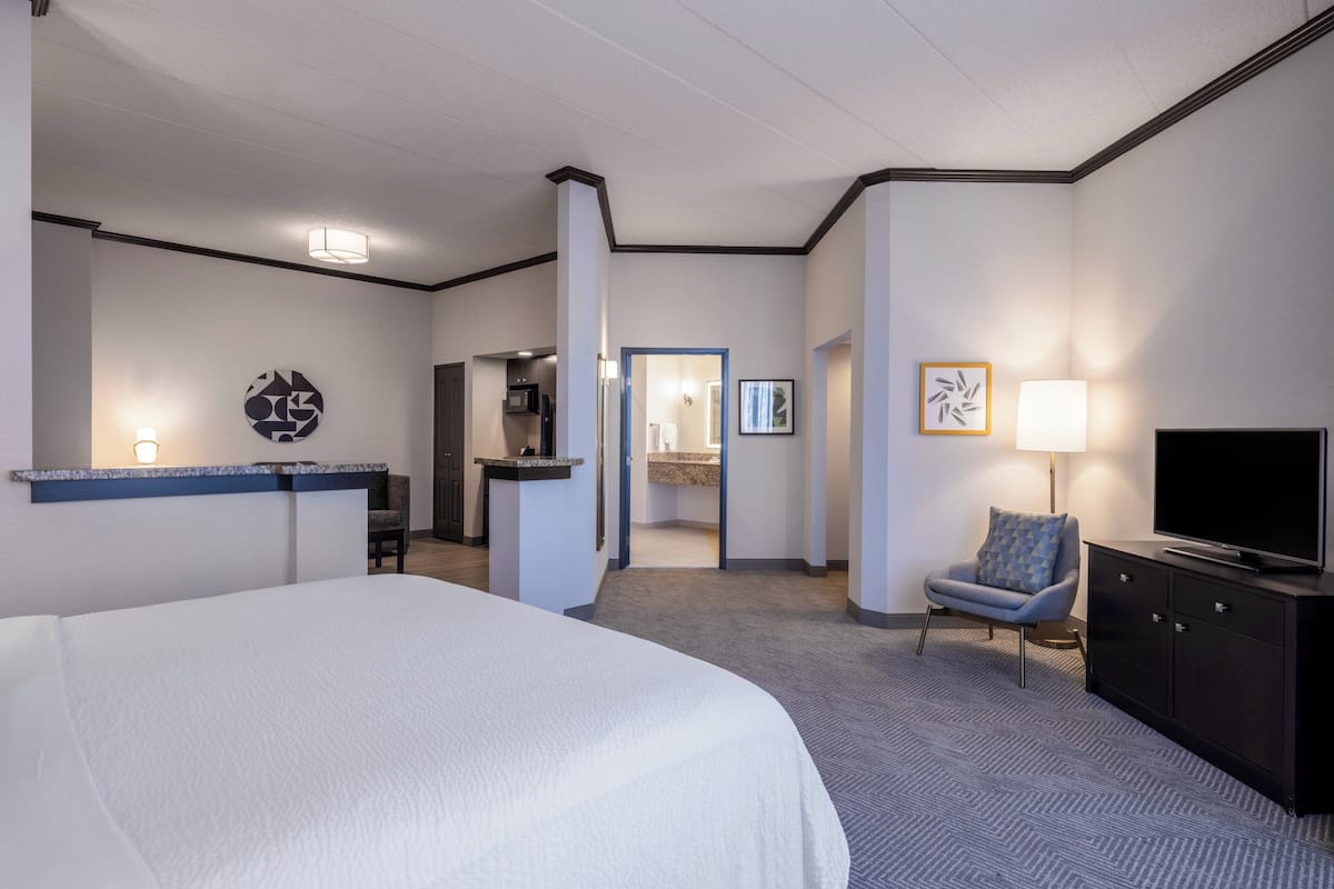 A hotel room with a large bed, a desk with a chair, a TV, a chair with a cushion, and a bathroom visible through an open door.