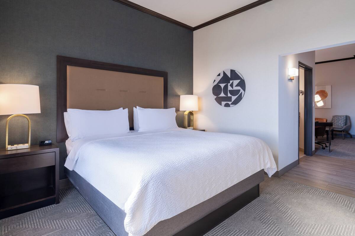 A hotel room with a large bed, two bedside tables with lamps, a wall art piece, and a view into another room.