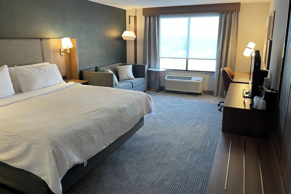 A hotel room with a large bed, a desk with a computer, a chair, a sofa, a window with curtains, and an air conditioning unit.