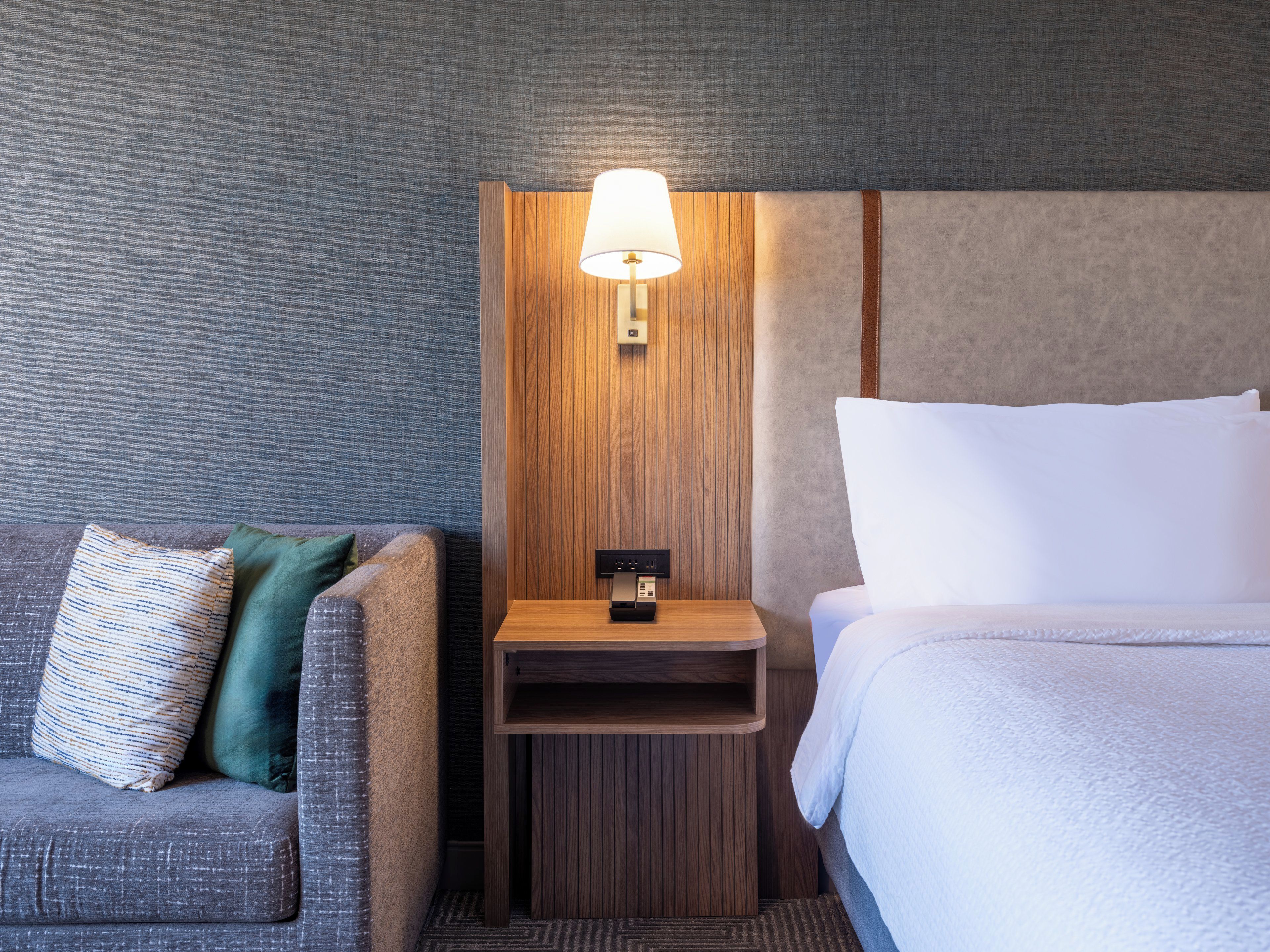 Ett hotellrum med en grå soffa, ett träbord vid sängen med en lampa och en säng med vita sängkläder.