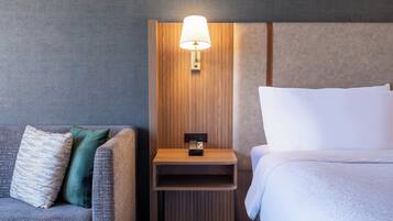 Ett hotellrum med en grå soffa, ett träbord vid sängen med en lampa och en säng med vita sängkläder.