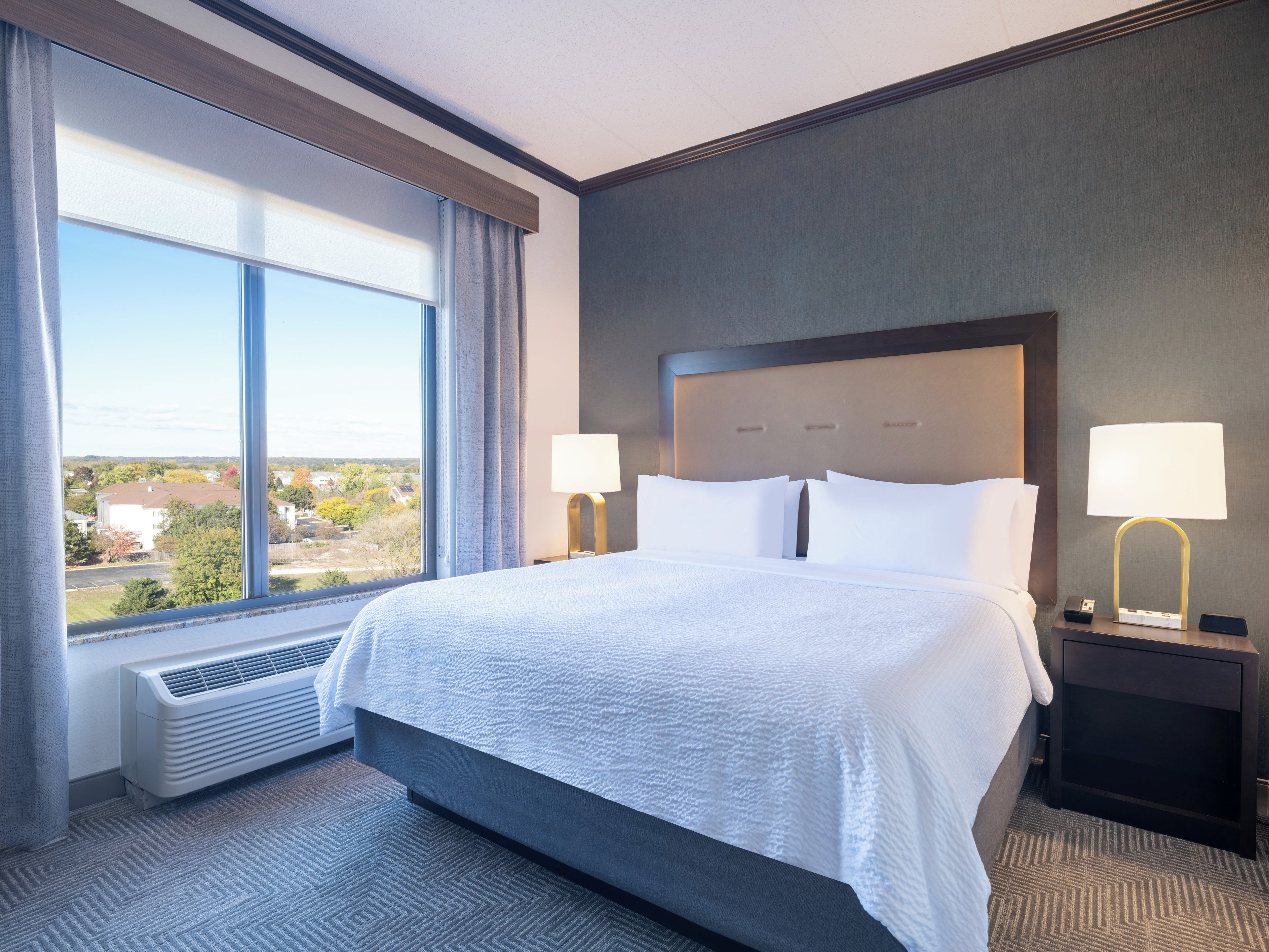 A hotel room with a large bed, two bedside tables with lamps, a window with curtains, and a view of the outside.