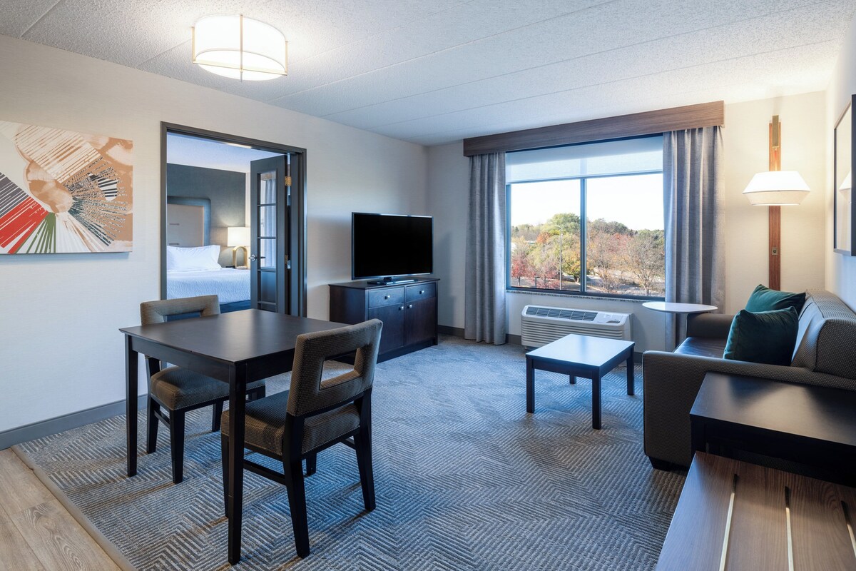 A hotel room with a dining area, a sofa, a TV, and a large window with a view of trees.