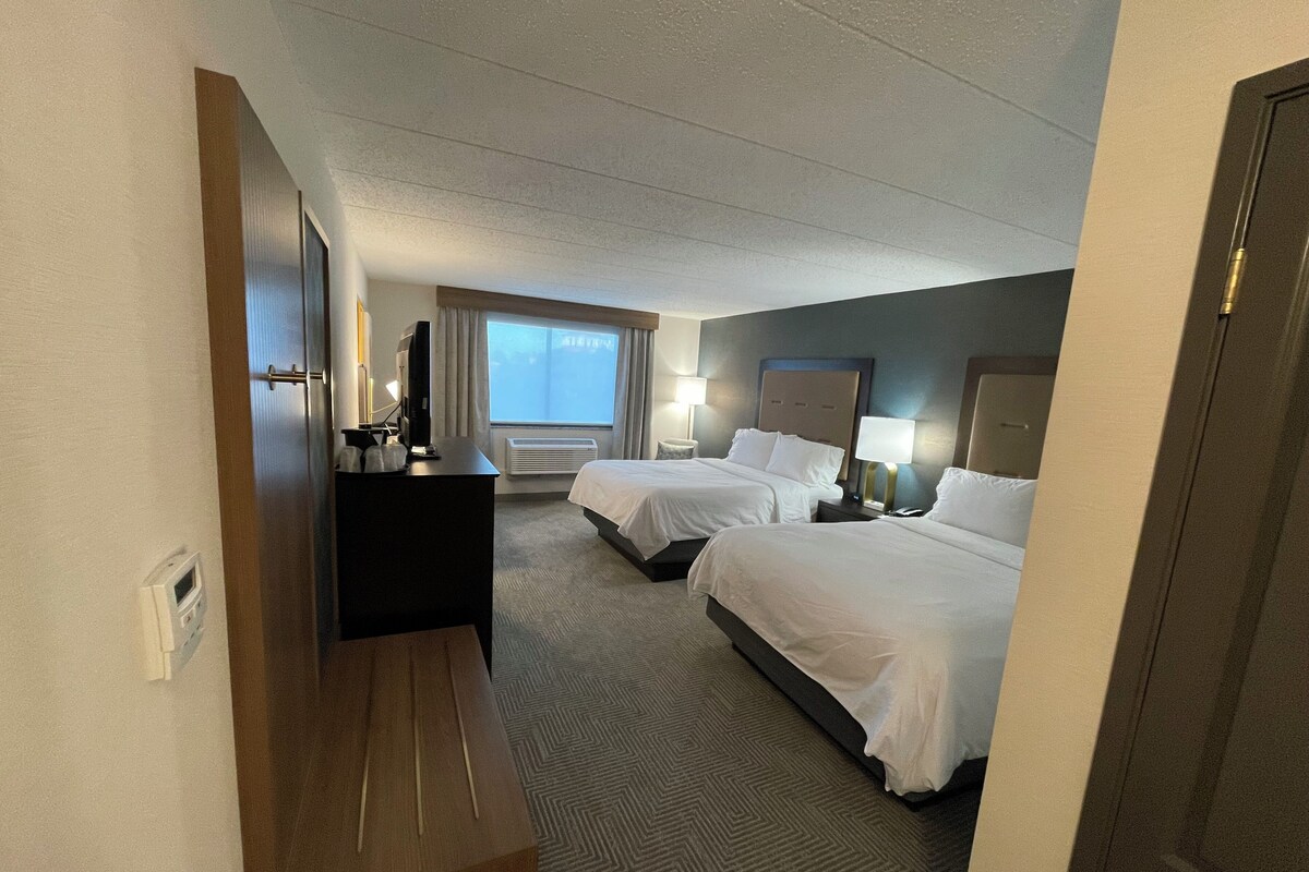A hotel room with two beds, a desk with a computer, a window with curtains, and a wall-mounted air conditioner.