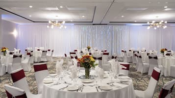 A banquet hall with round tables set for dining, white chairs with red sashes, and a backdrop of white curtains with lights.