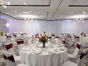 A banquet hall with round tables set for dining, white chairs with red sashes, and a backdrop of white curtains with lights. - Holiday Inn Chicago Northwest-Elgin by IHG (Elgin)