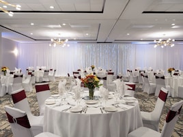 Un salón de banquetes con mesas redondas dispuestas para cenar, sillas blancas con cintas rojas y un telón de fondo de cortinas blancas con luces.