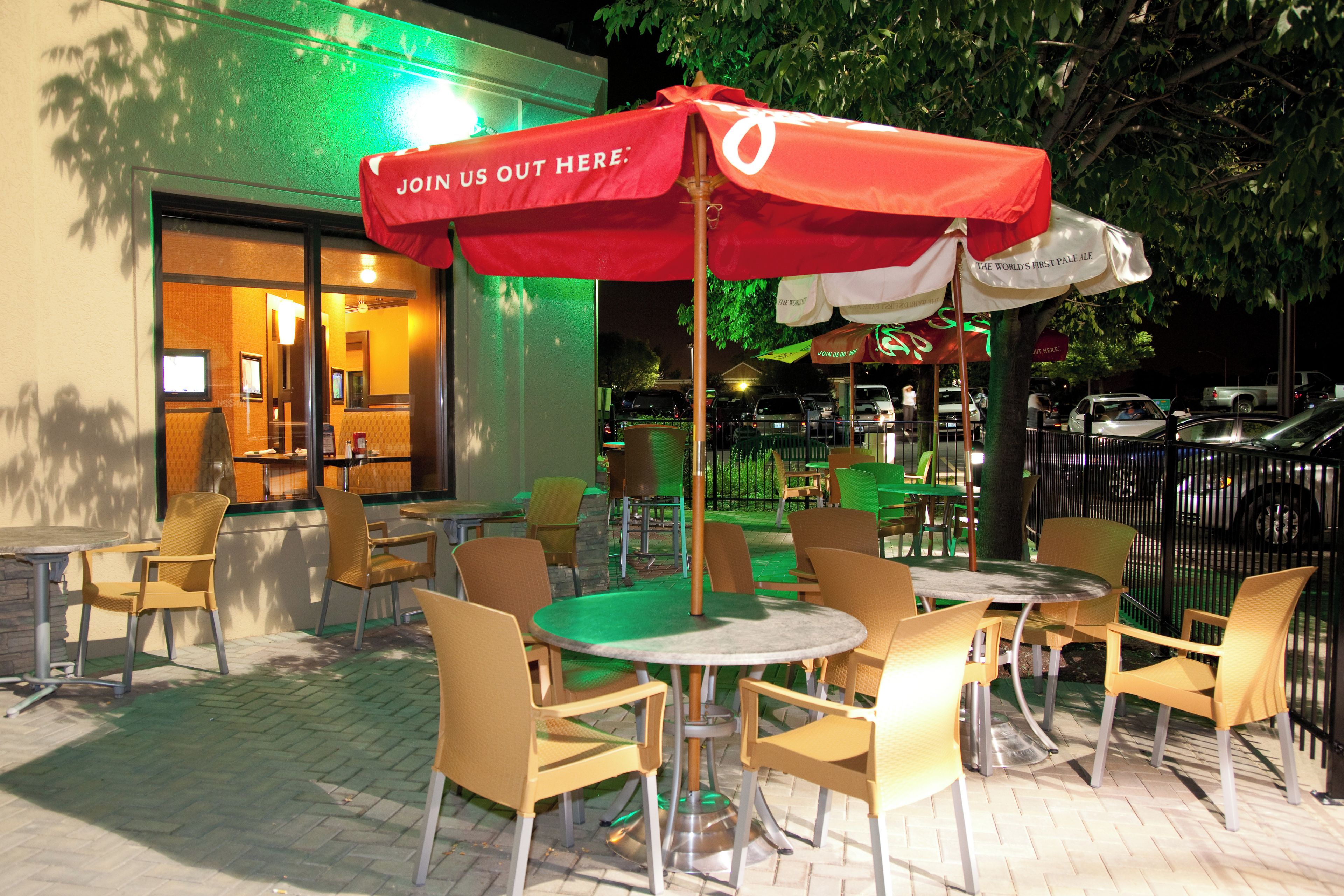 An outdoor seating area with tables and chairs, a red umbrella, and a building entrance.