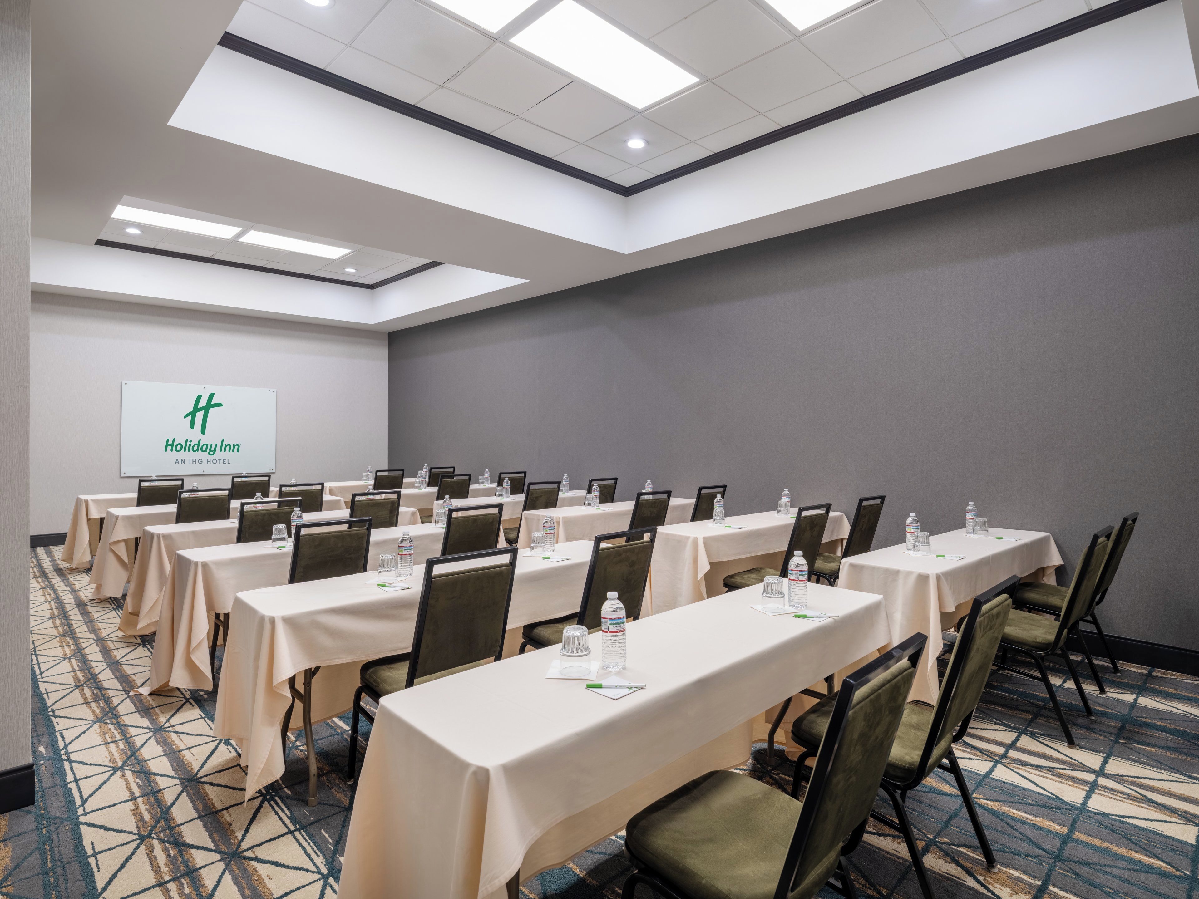 Un salón de conferencias con varias mesas y sillas, una pantalla de proyector mostrando el logotipo de Holiday Inn y una alfombra estampada.