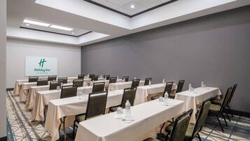 Un salón de conferencias con varias mesas y sillas, una pantalla de proyector mostrando el logotipo de Holiday Inn y una alfombra estampada.