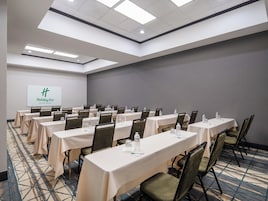 Un salón de conferencias con varias mesas y sillas, una pantalla de proyector mostrando el logotipo de Holiday Inn y una alfombra estampada.