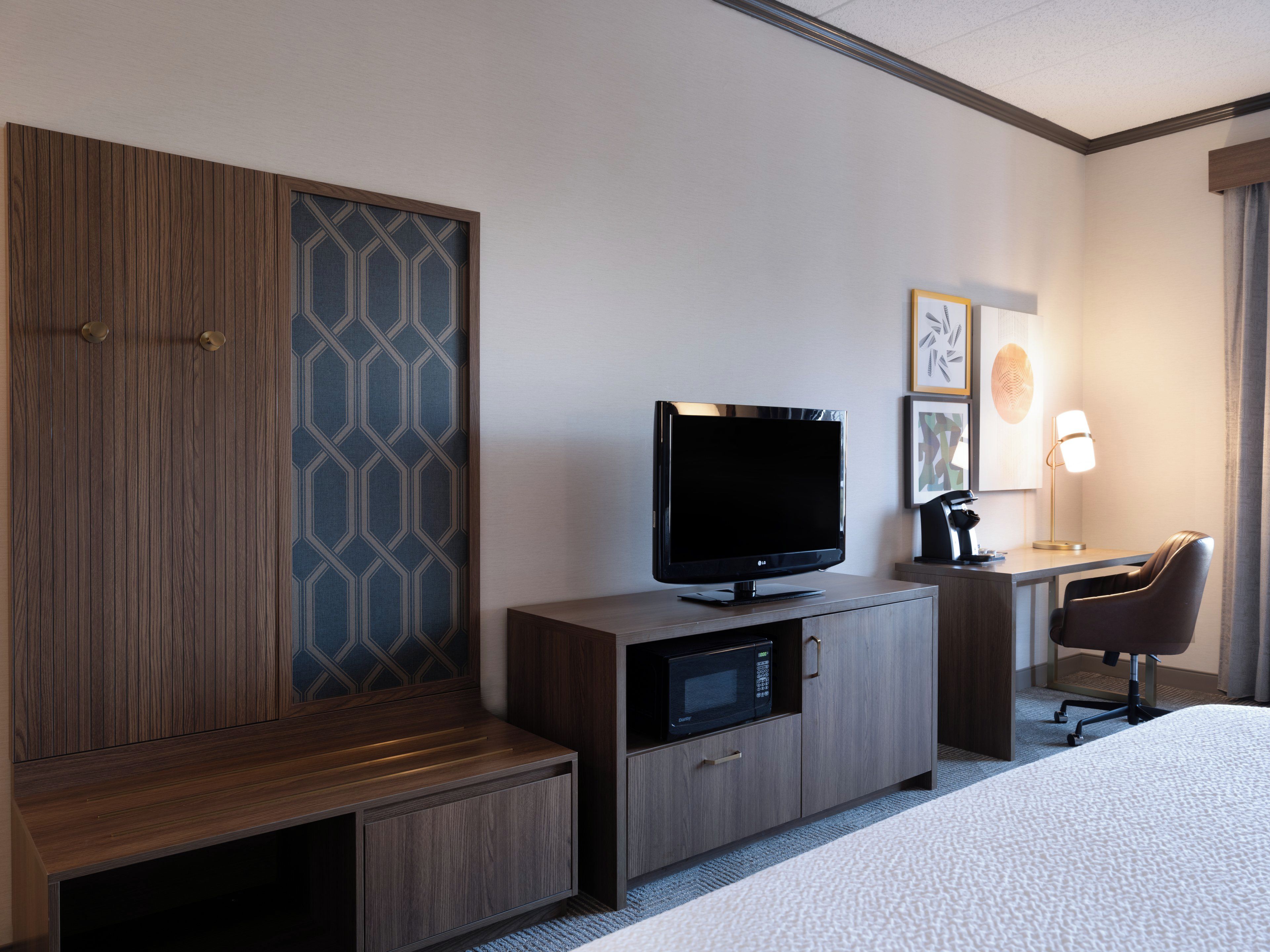 Quarto de hotel com uma cama, uma televisão sobre um móvel de madeira, uma escrivaninha com abajur e uma cadeira.
