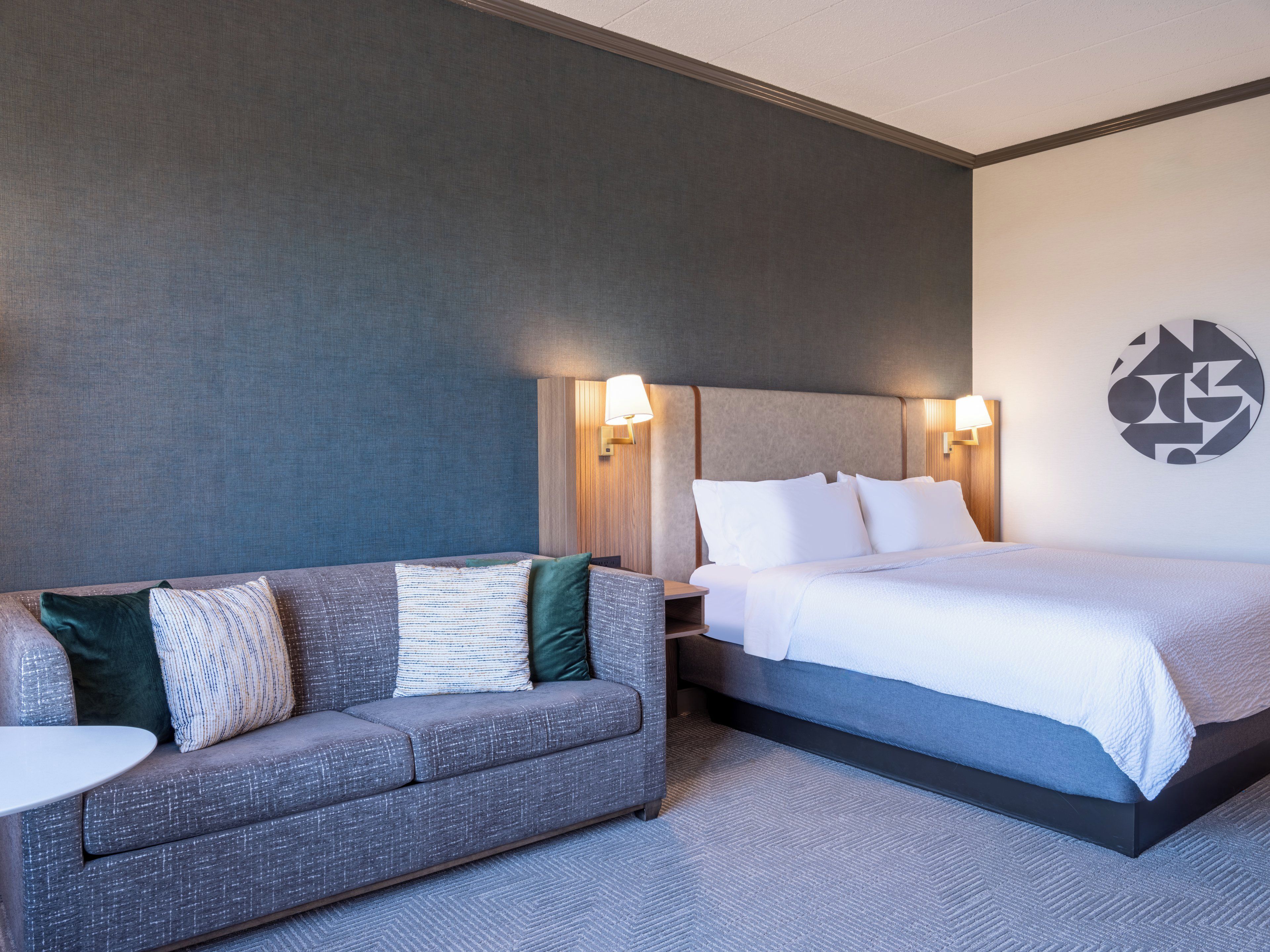 A hotel room with a grey sofa, a bed with white bedding, and a wall with a geometric design.