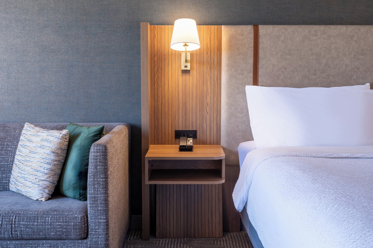 A hotel room with a grey sofa, a wooden nightstand with a lamp, and a bed with white bedding.