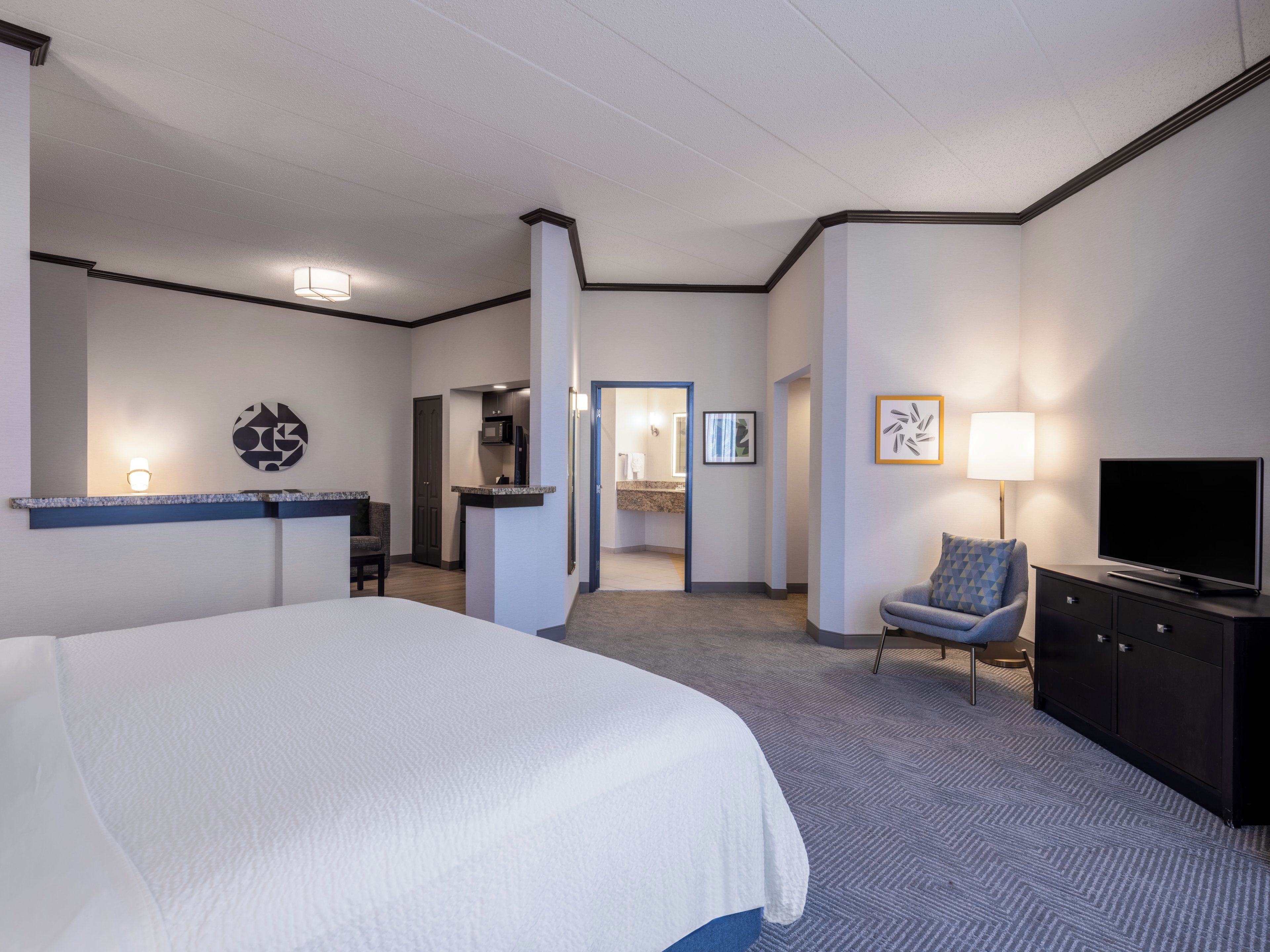 Camera d'albergo con un letto grande, una scrivania con una sedia, una televisione, una poltrona con un cuscino e un bagno visibile attraverso una porta aperta.