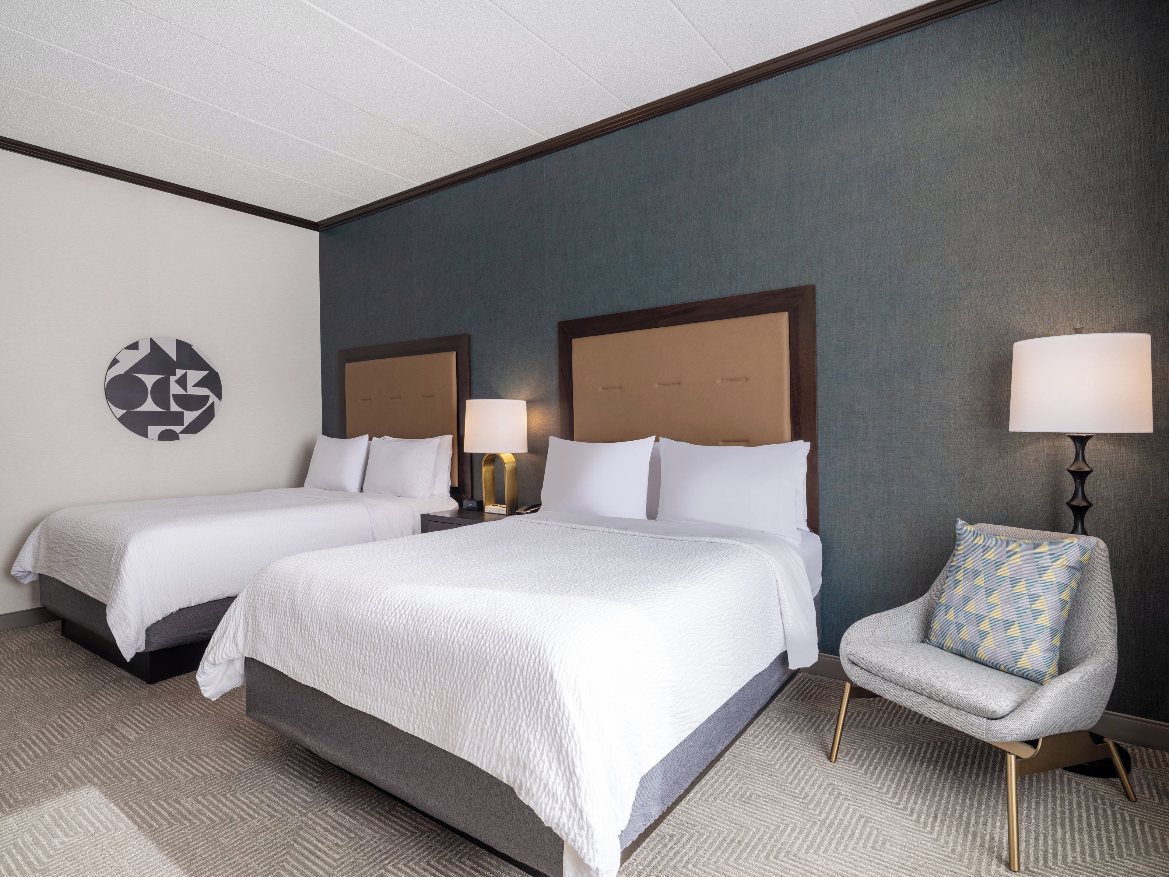 A hotel room with two beds, a chair, a lamp, and a wall art piece.