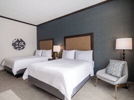 Habitación de hotel con dos camas, una silla, una lámpara y una obra de arte en la pared.