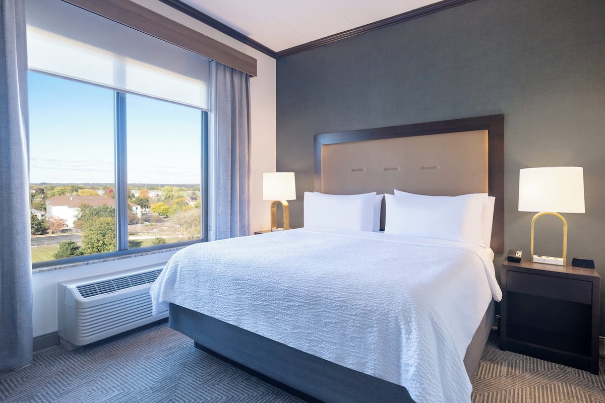 A hotel room with a large bed, two bedside lamps, a window with curtains, and a view of the outside.