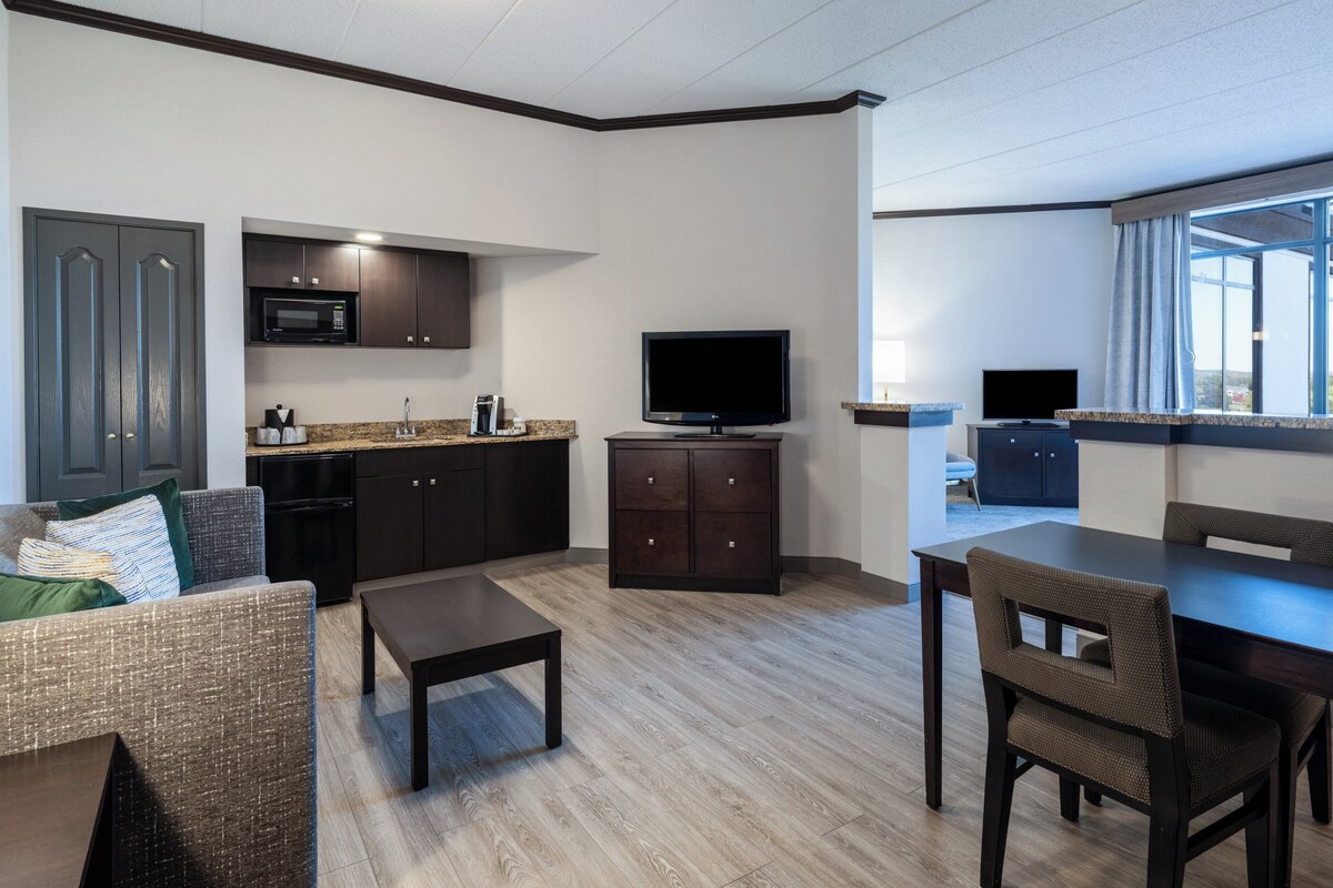 A modern hotel room with a kitchenette, a flat-screen TV, a dining area with a table and chairs, and a large window offering a view.