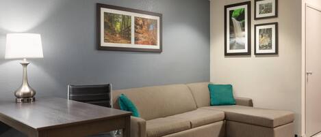 A room with a beige sectional sofa, a small table with a lamp, and a chair. There are framed pictures on the wall.