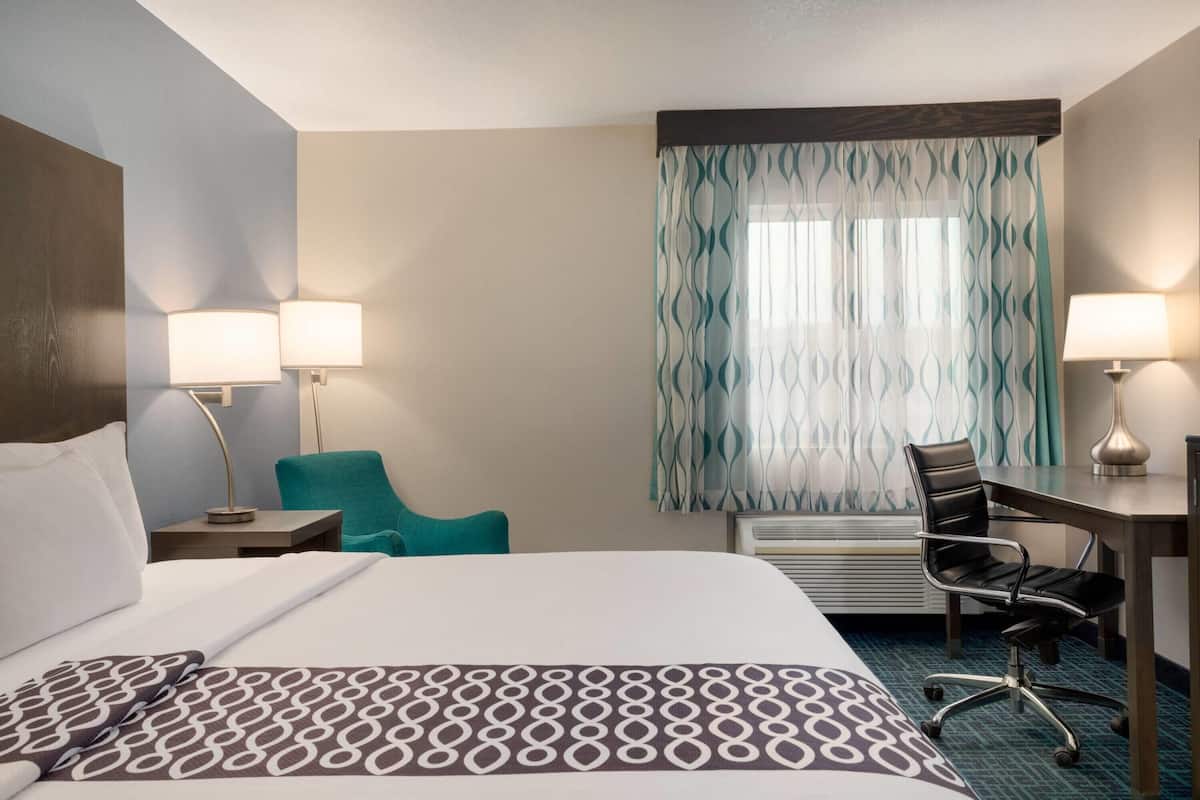 Hotelkamer met een groot bed, twee bedlampen, een bureau met stoel en een raam met gordijnen met dessin.