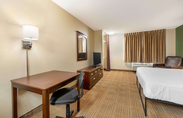 A hotel room with a bed, a desk with a chair, a television on a dresser, a mirror, and a window with curtains.