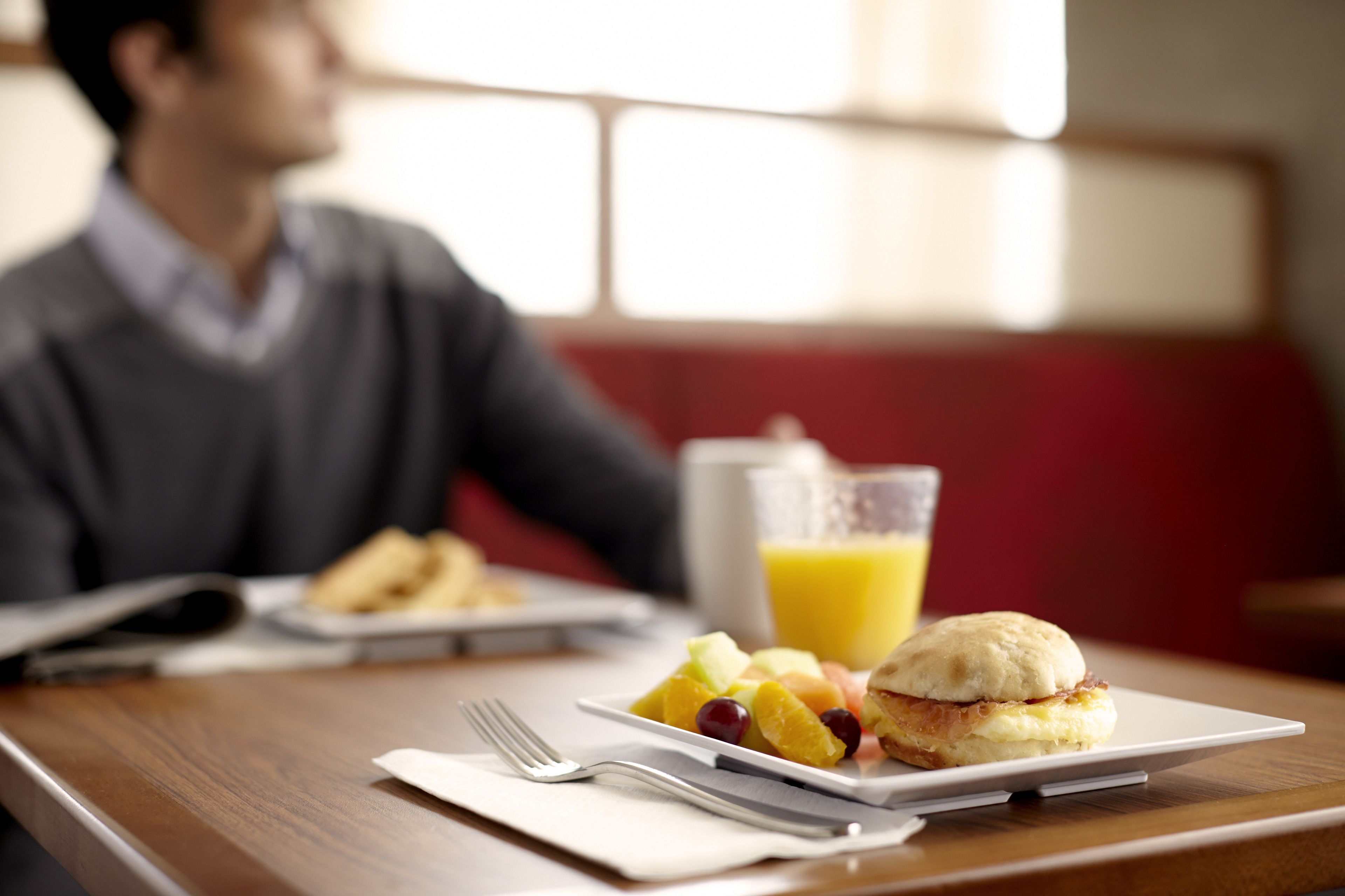 Une table en bois dressée pour le petit-déjeuner, avec une assiette de fruits, un sandwich et un verre de jus d’orange.