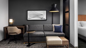 Een moderne hotelkamer met een grijze bank, een klein rond tafeltje, een bureau met stoel, een vloerlamp en een abstract kunstwerk in een lijst aan de muur.