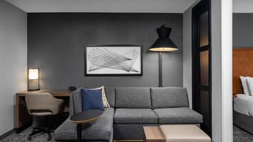 A modern hotel room with a grey sofa, a small round table, a desk with a chair, a lamp, and a framed abstract artwork on the wall.