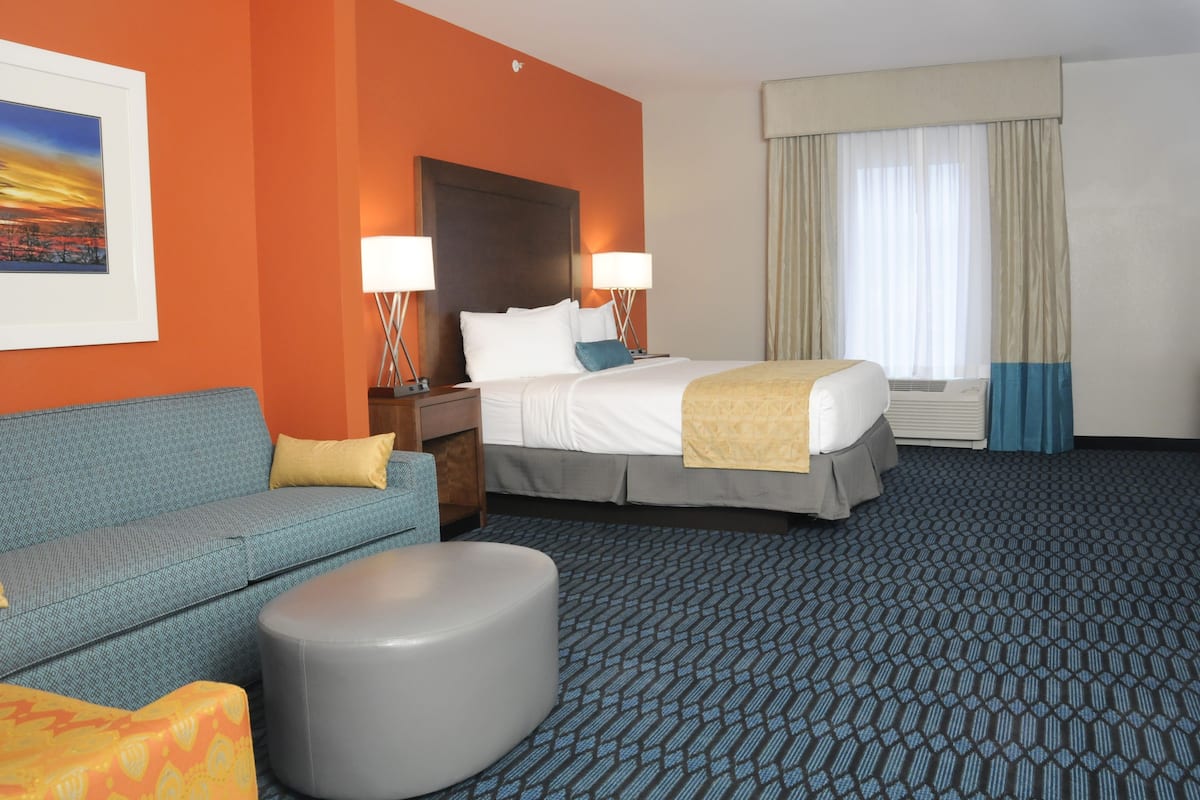 A hotel room with a bed, a sofa, a round ottoman, a nightstand, two lamps, a window with curtains, and a framed picture on the wall.