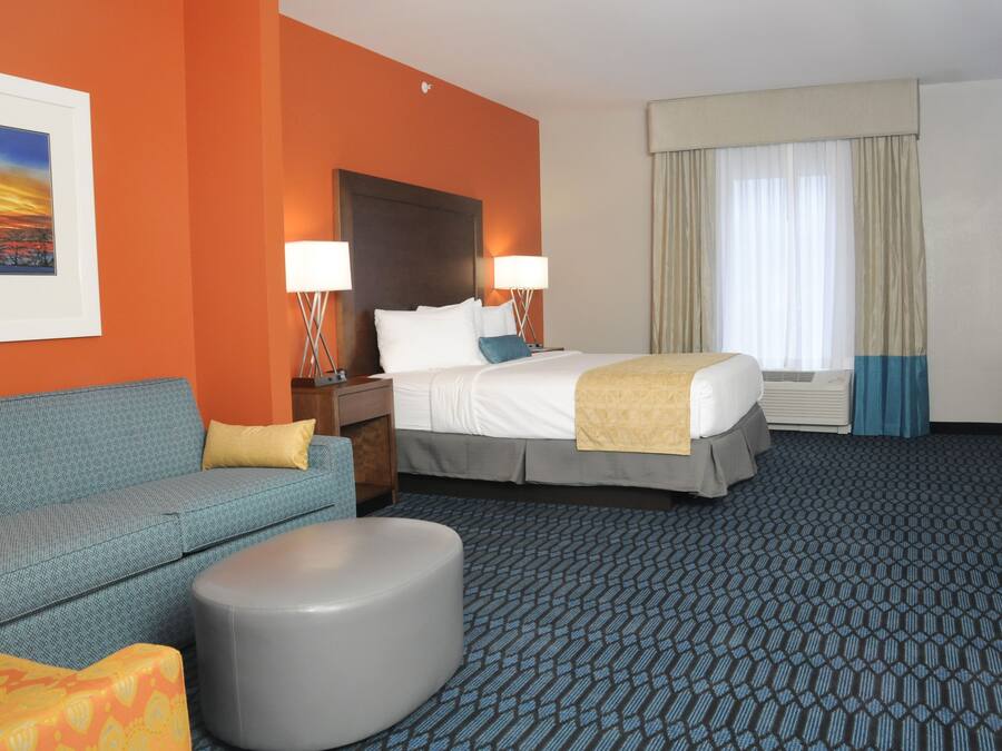 A hotel room with a bed, a sofa, a round ottoman, a nightstand, two lamps, a window with curtains, and a framed picture on the wall.