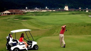 A golf course with a clubhouse, a golf cart, and a person preparing to hit a golf ball.