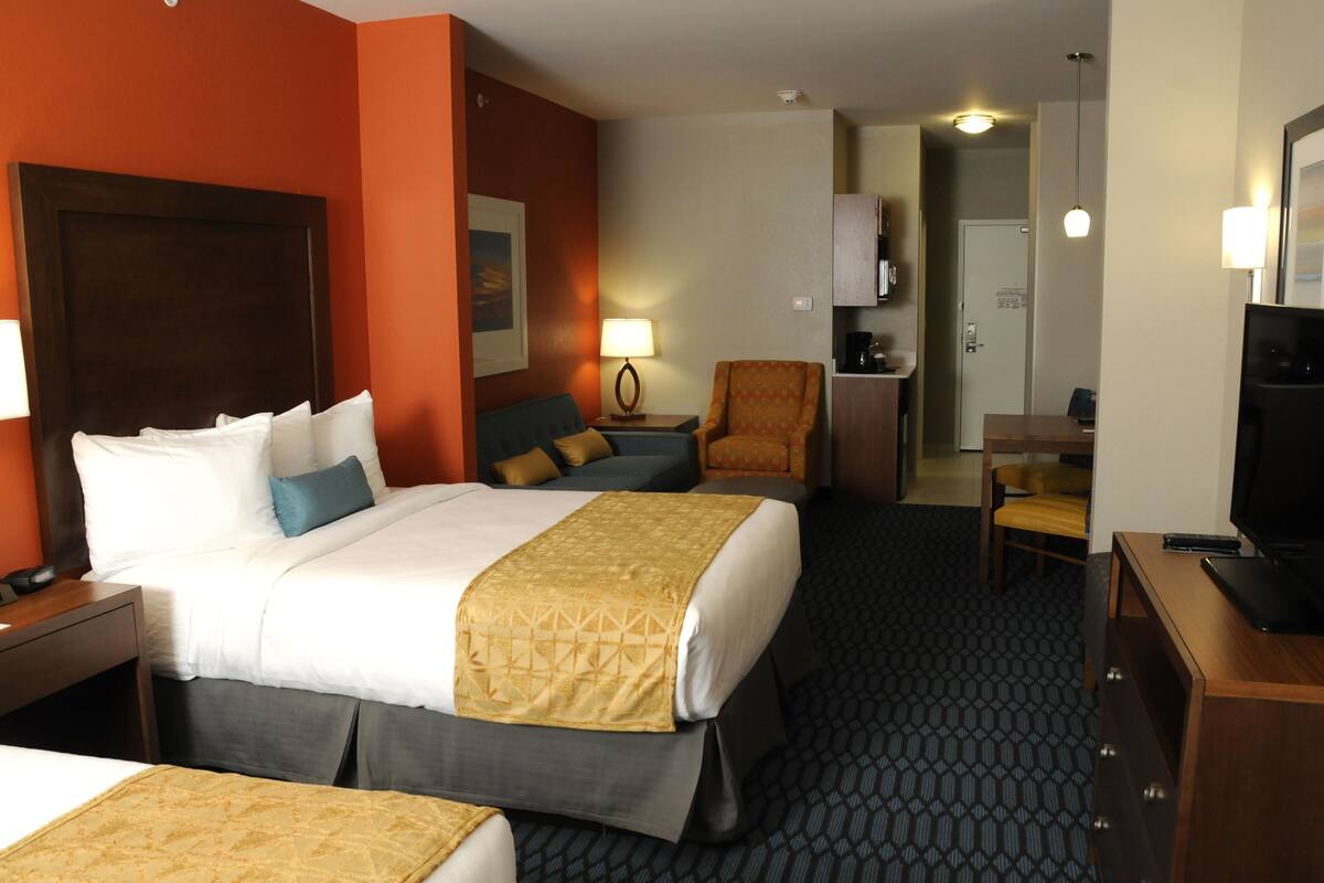 A hotel room with a large bed, a smaller bed, a desk with a chair, a TV, and a lamp.