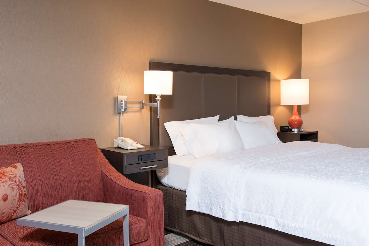 A hotel room with a bed, a red armchair, a small table, and two bedside lamps.