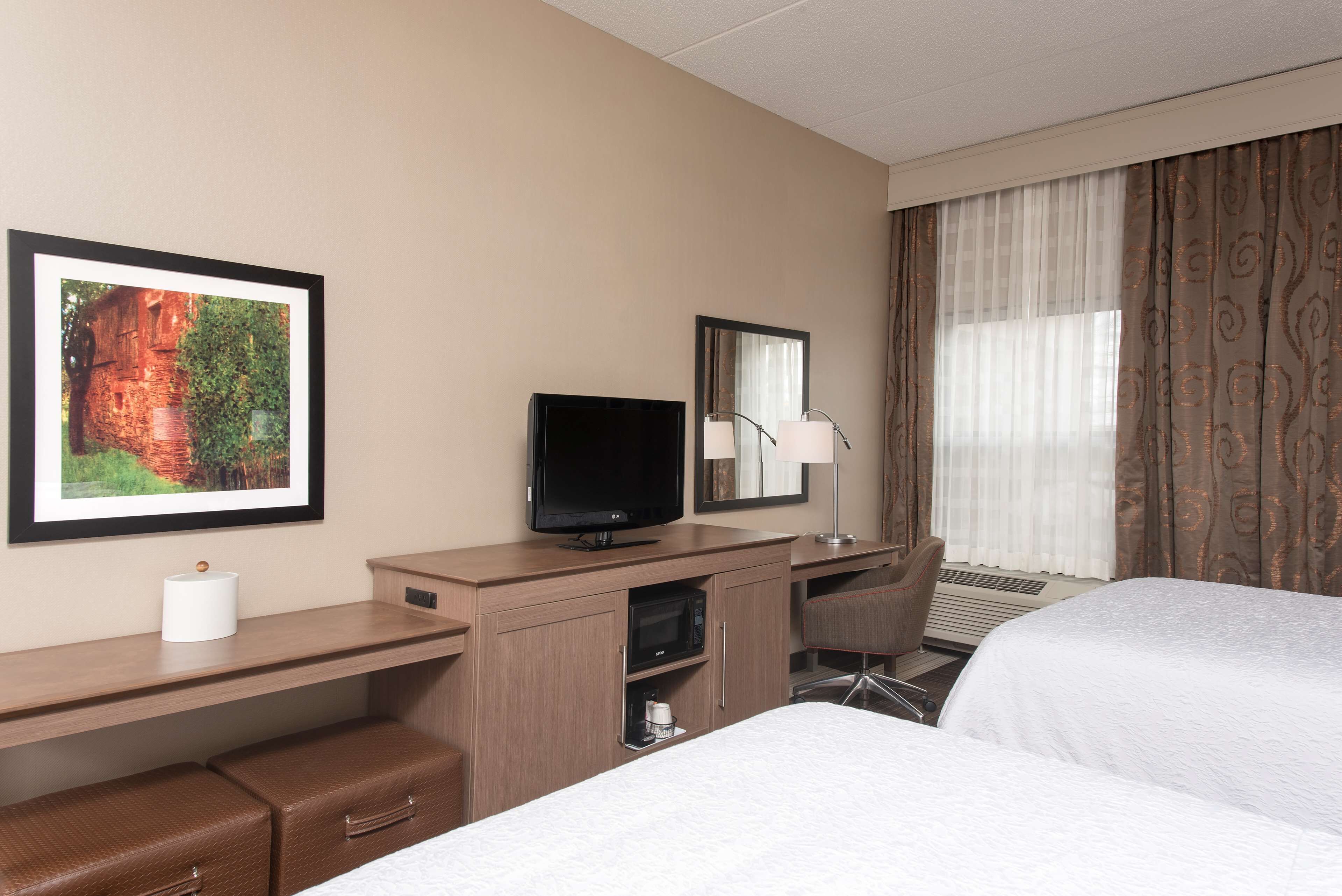 A hotel room with a bed, a desk with a television, a mirror, a lamp, and a picture on the wall.