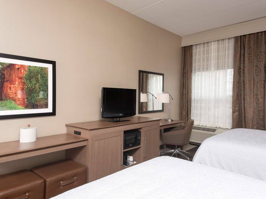 A hotel room with a bed, a desk with a television, a mirror, a lamp, and a picture on the wall.