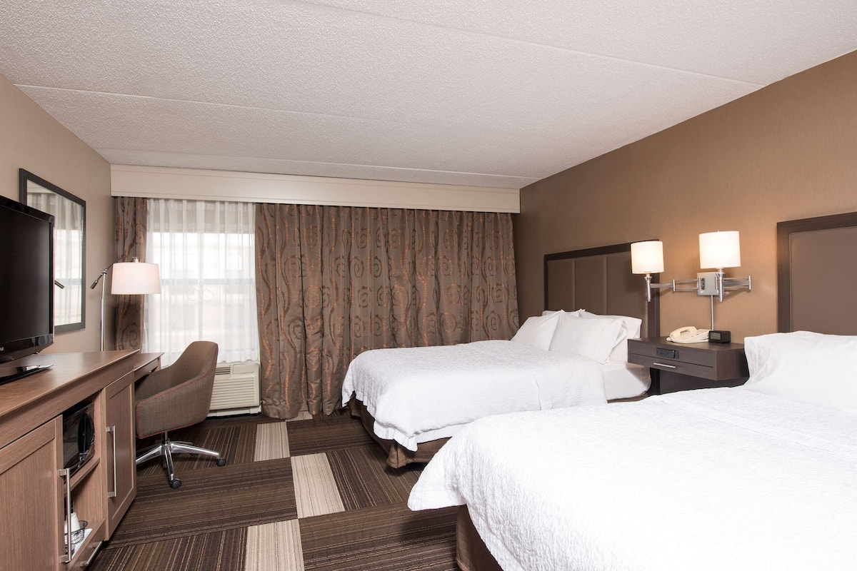 A hotel room with two beds, a desk with a TV, a chair, and a desk lamp.