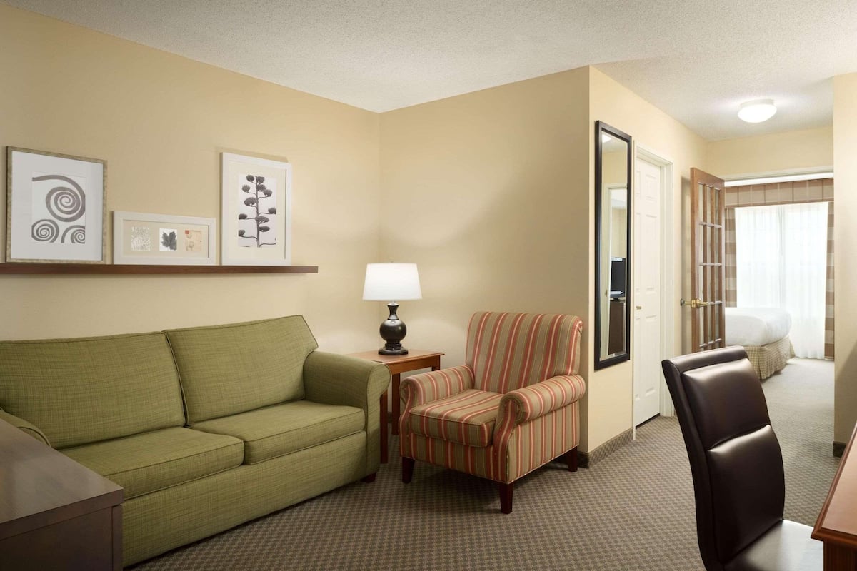 A hotel room with a green sofa, a striped armchair, a black leather chair, a small table with a lamp, and framed artwork on the wall.