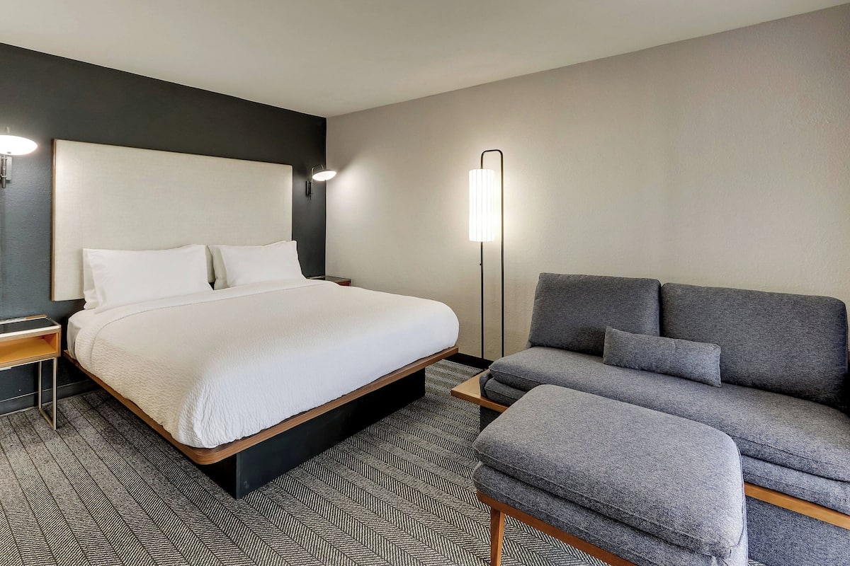 Una habitación de hotel moderna con una cama grande, un sofá gris y una mesita de noche.