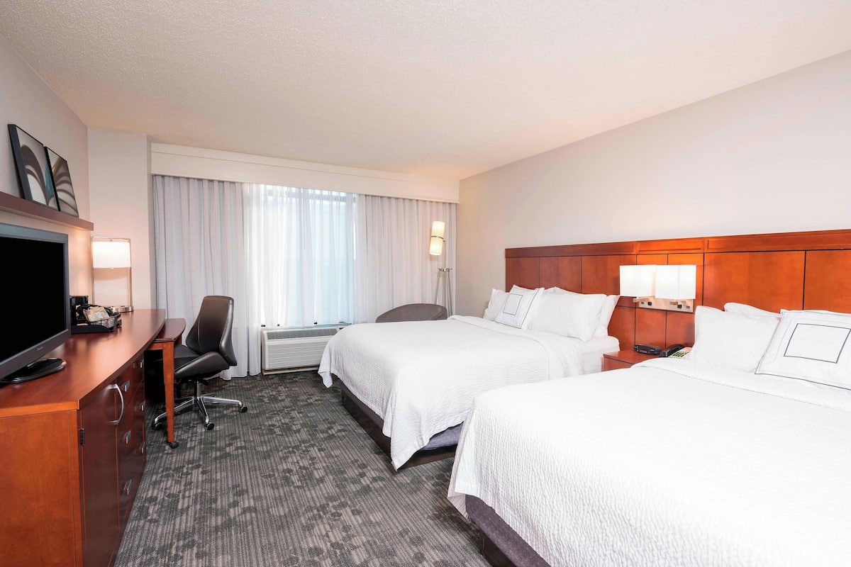 A hotel room with two beds, a desk with a TV, a chair, and a lamp.