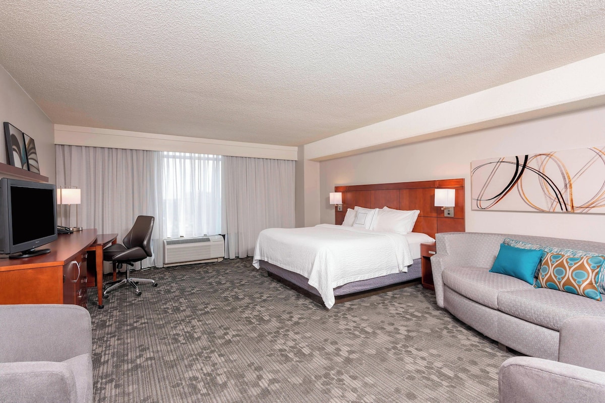 A hotel room with a bed, a sofa, a desk with a TV, and a large abstract painting on the wall.
