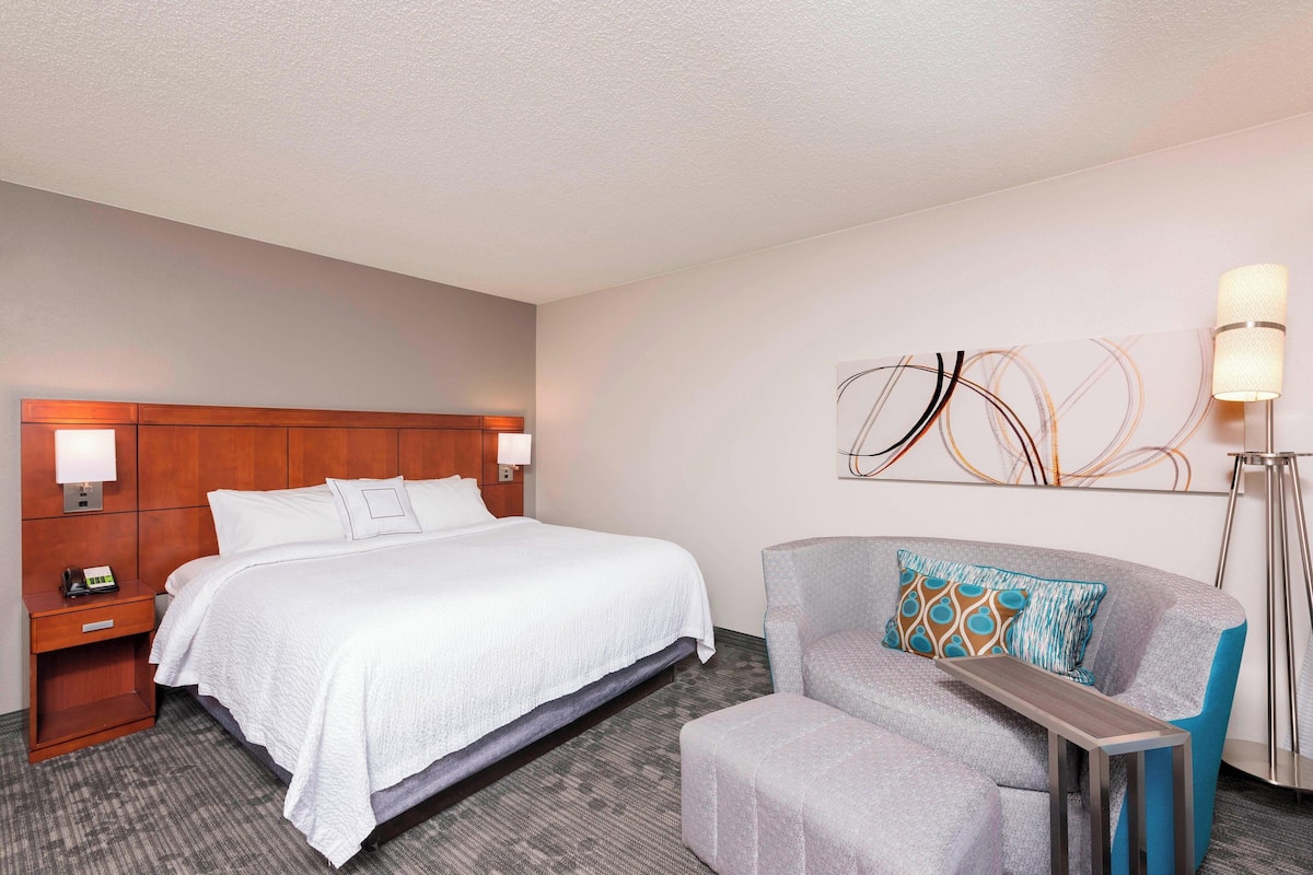 A hotel room with a large bed, a nightstand with a phone, a gray armchair with a patterned pillow, and a modern abstract wall art piece.