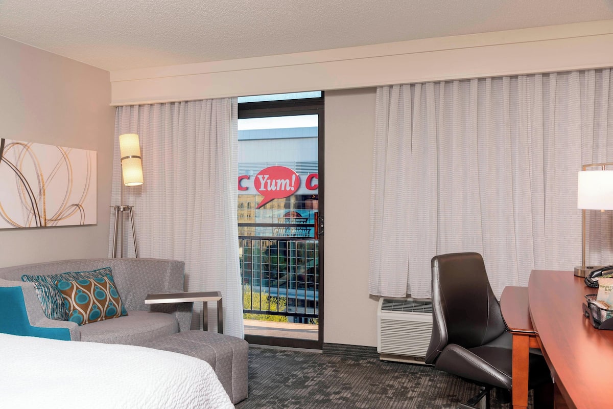 A hotel room with a gray sofa, a small table, a desk, a chair, and a large window with a view of a building and a sign that says "C Yum!".
