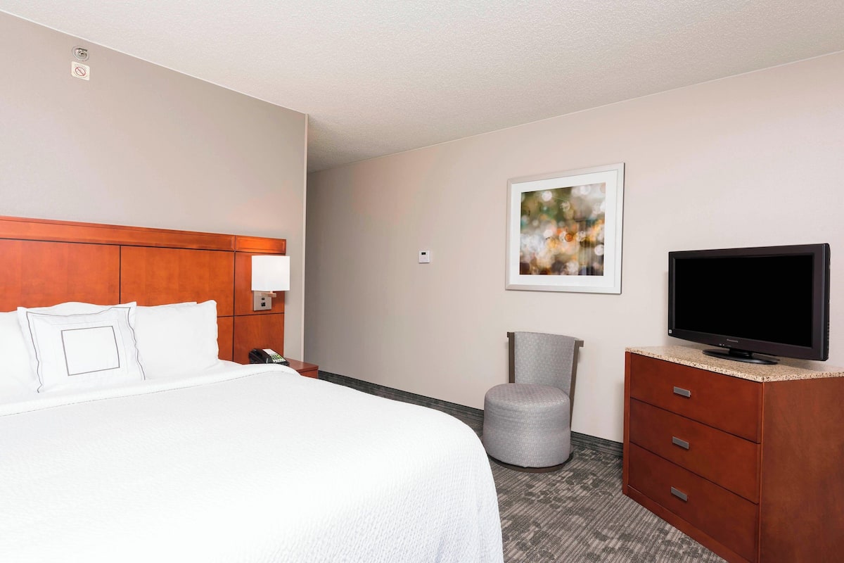 A hotel room with a bed, a television on a wooden dresser, a gray ottoman, and a framed picture on the wall.