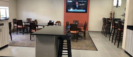 A conference room with a large screen displaying a news broadcast, multiple tables and chairs, and a bar area with stools.