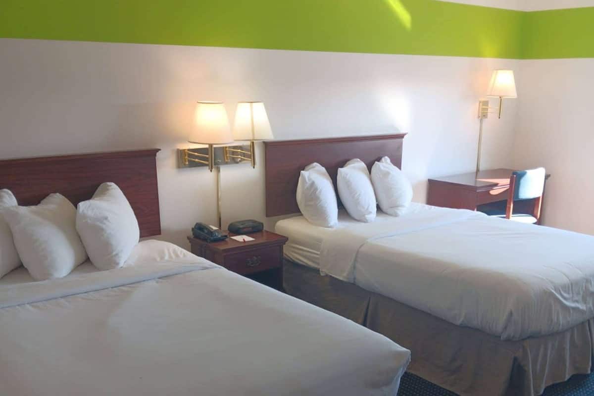 A hotel room with two beds, each with white bedding and pillows, a wooden headboard, bedside tables with lamps, and a small desk with a chair.