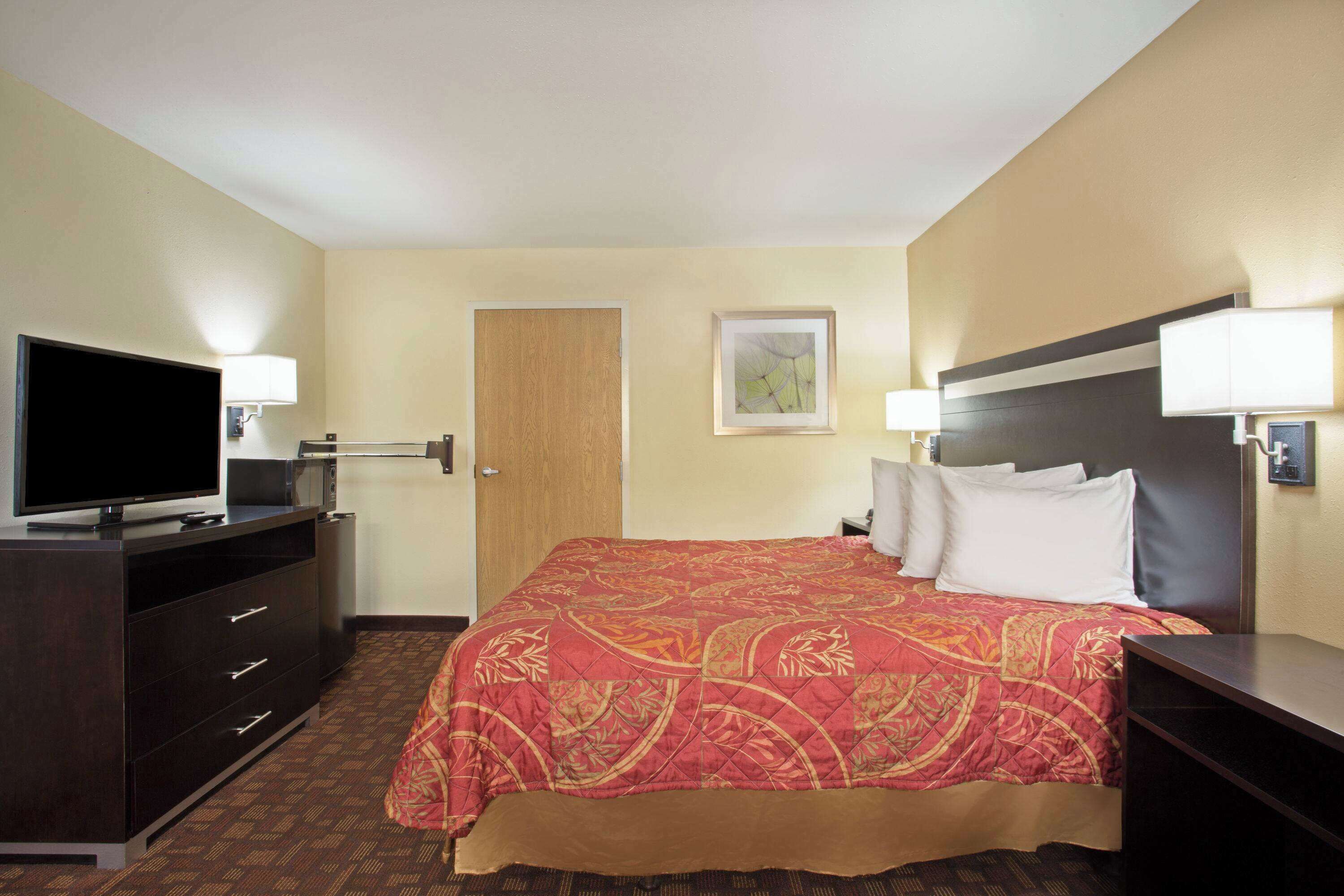 A hotel room with a large bed, a television on a dresser, and two wall-mounted lamps.