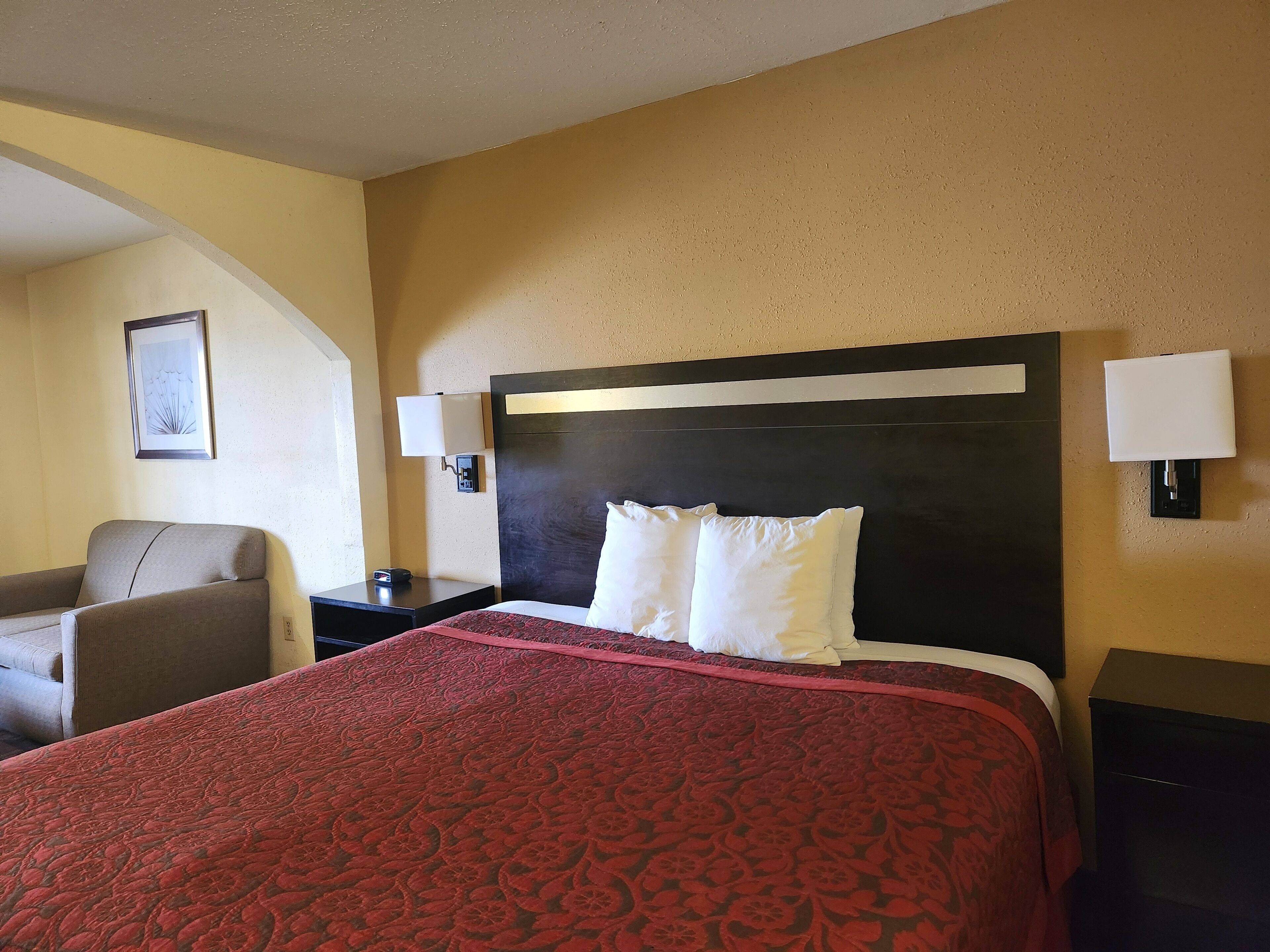 Habitación de hotel con una cama grande, una mesita de noche, una lámpara, una silla y un cuadro en la pared.