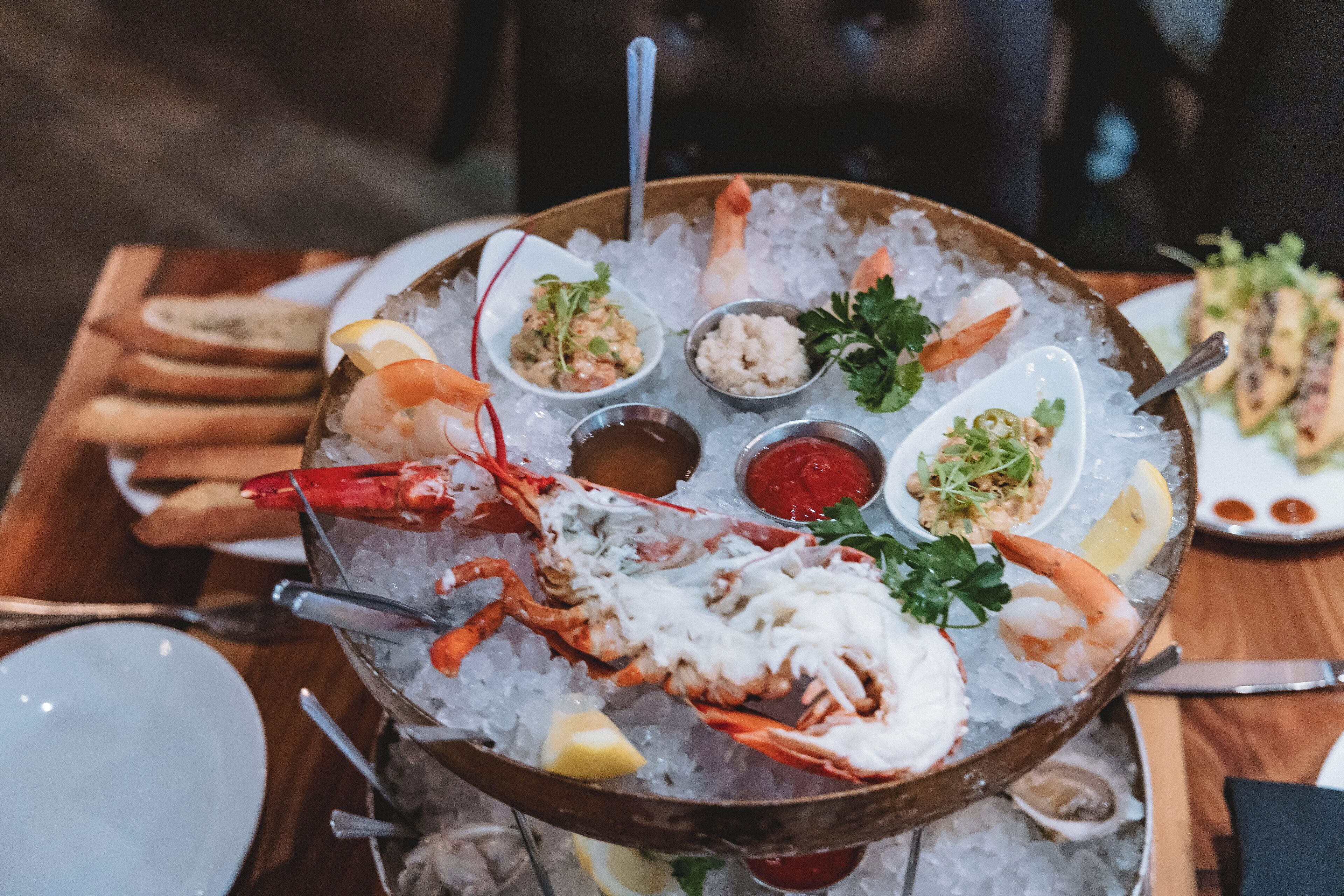 Un plato de mariscos sobre hielo con diversas preparaciones, incluyendo camarones, langosta y otros mariscos, acompañado de gajos de limón y adornos.