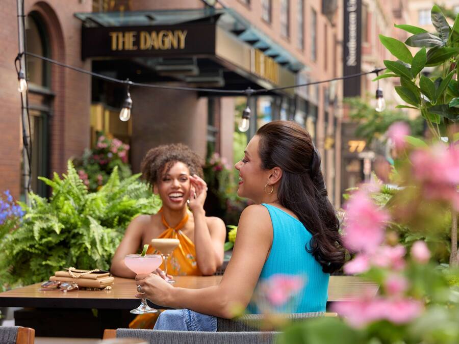 Two people sitting at an outdoor table with a drink, surrounded by greenery and a building with a sign reading "THE DAGNY".